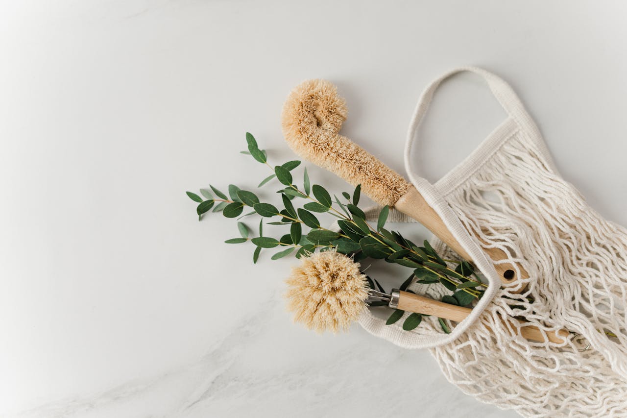 hero-img-01 Eco-friendly cleaning brushes and eucalyptus branches in a reusable net bag on white surface.